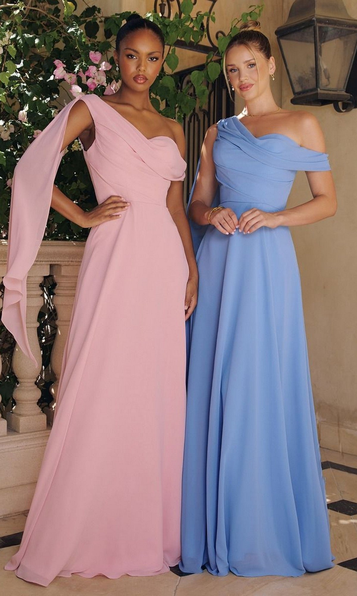 Two women showcase the Amoris T716 Asymmetric Long A-Line Prom Dress—one in pink with a draped sleeve, the other in light blue—posing outdoors by a stone railing and lush greenery, highlighting chic affordability.